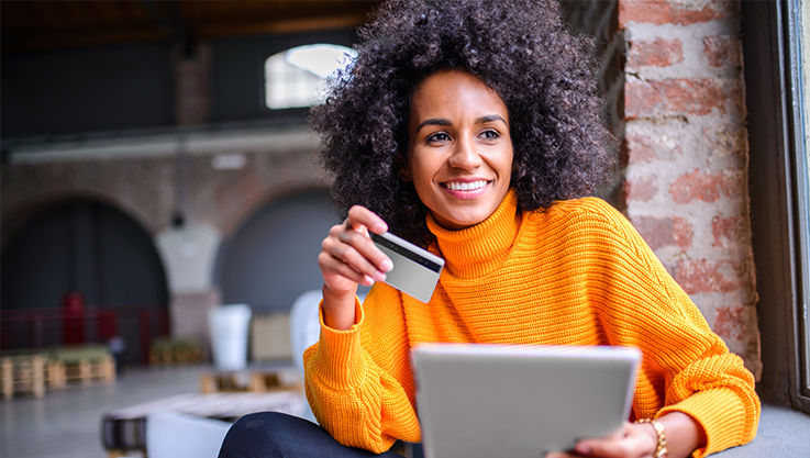 A woman wearing a bright orange sweater sits at a computer holding a gold credit card. 