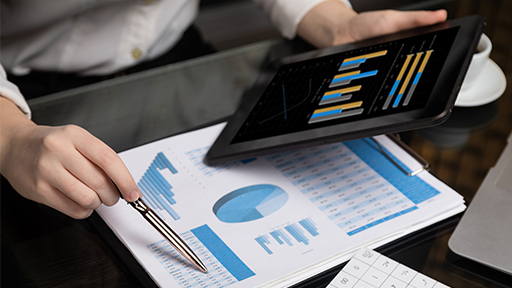 Looking down on a desk where a person is holding a tablet over a table with papers. The tablet and papers both show business graphs. 