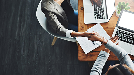 Two professionals sit at a corner of a desk with laptops, a notebook and a small green plant. They are shaking hands.