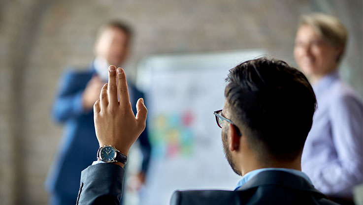 From behind, the photo captures a person in a suit raising their hand. In front are two individuals presenting.